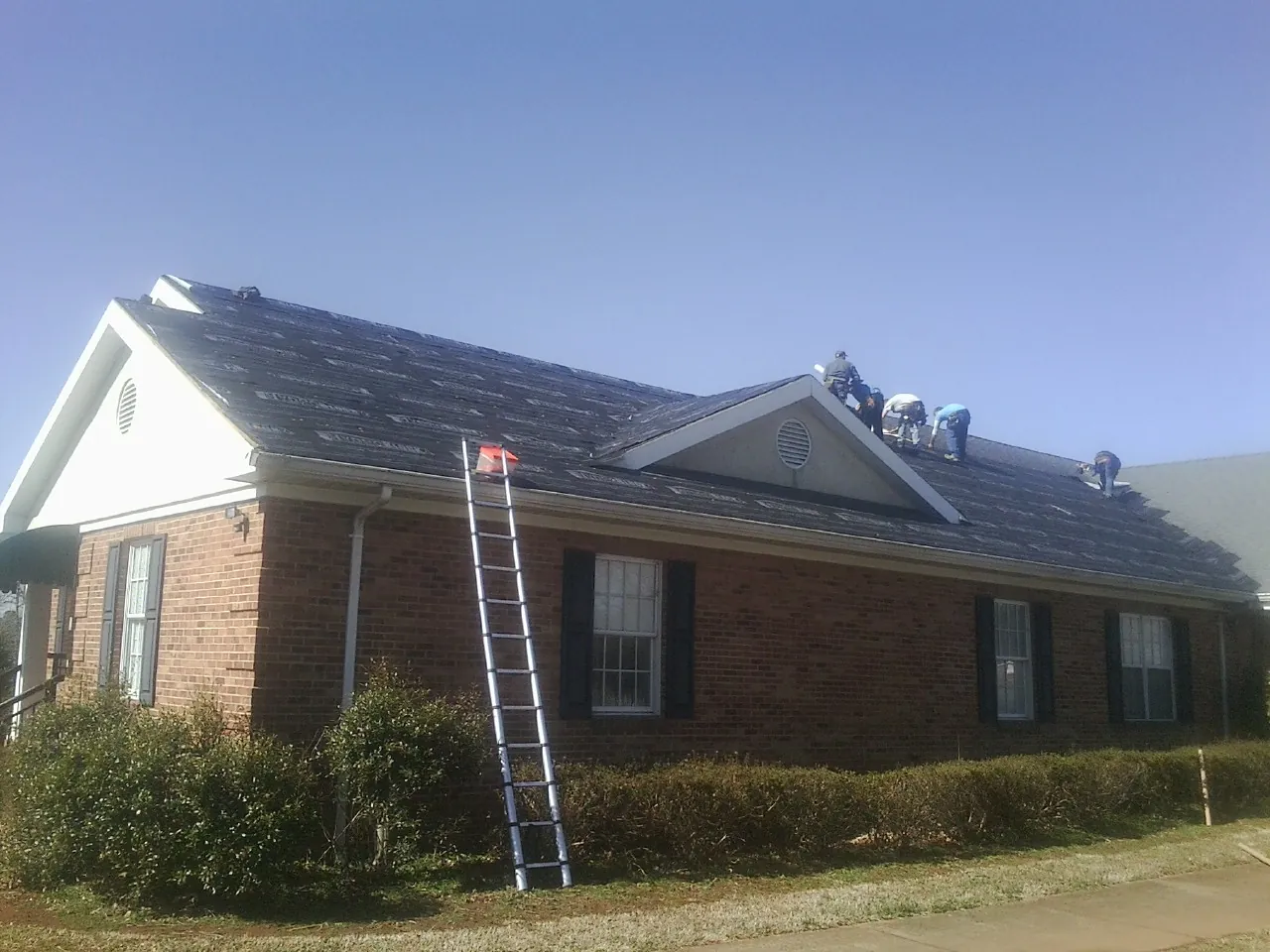Crew tearing shingles off a brick ranch roof with a ladder set — Emergency Roof Repair work by our crew in Roanoke Rapids