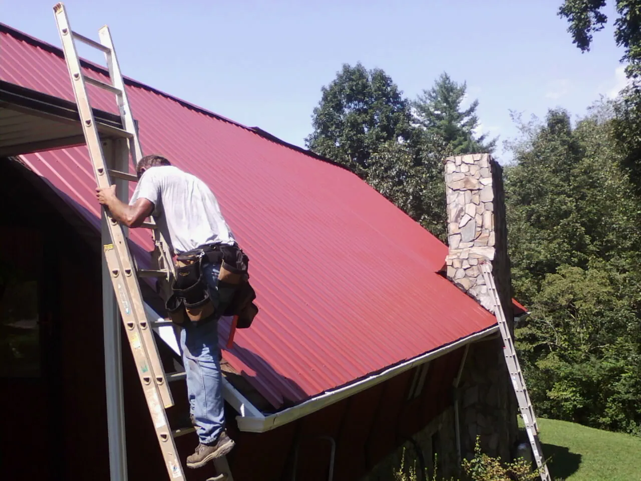 Roofer on a ladder beside a red metal roof and stone chimney — Soffit & Fascia Repair by Roofing Brothers in Roanoke Rapids